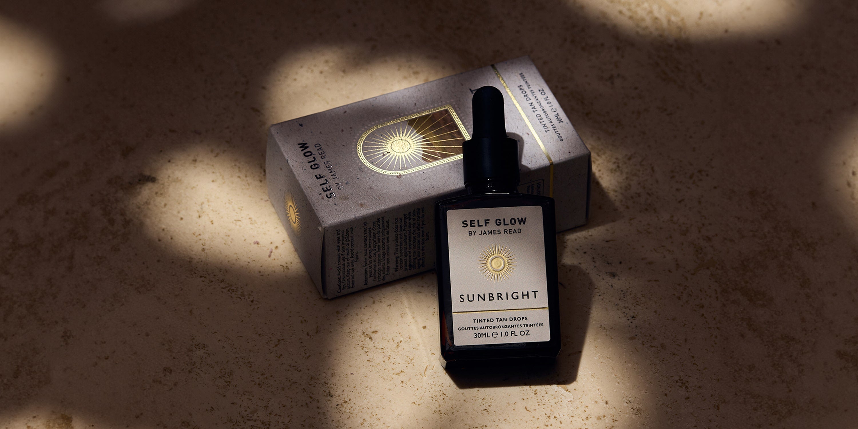 James Read's Self Glow skincare that glows: Sunbright Tinted Tan Drops in a recycled glass bottle leaning against recycled cardboard packaging, showcasing the brand's sustainability promise and commitment to eco-friendly materials, all under dappled light.