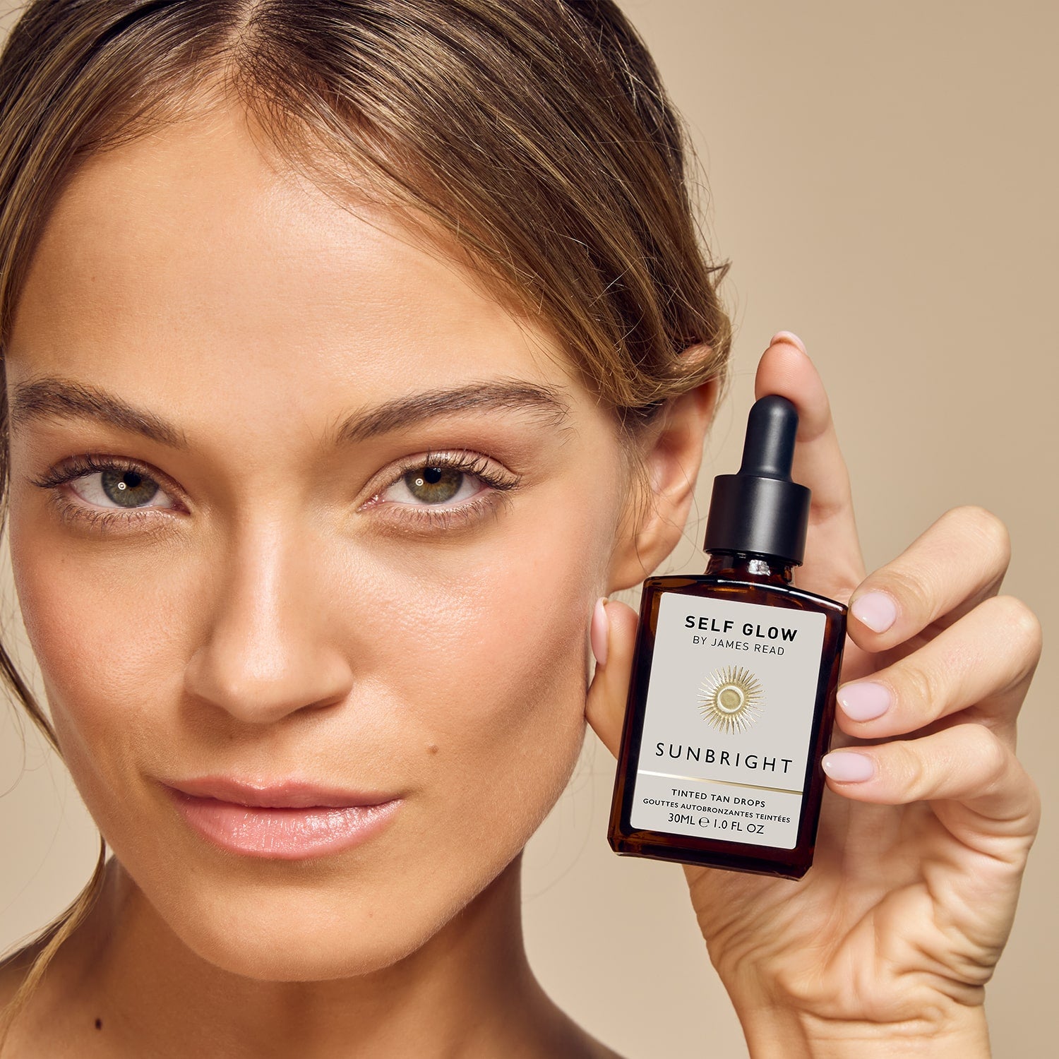 Discover a new way to glow with James Read's Self Glow skincare-infused sunless tanning Sunbright Tinted Tan Drops, held by a model for a natural-looking glow to the skin.