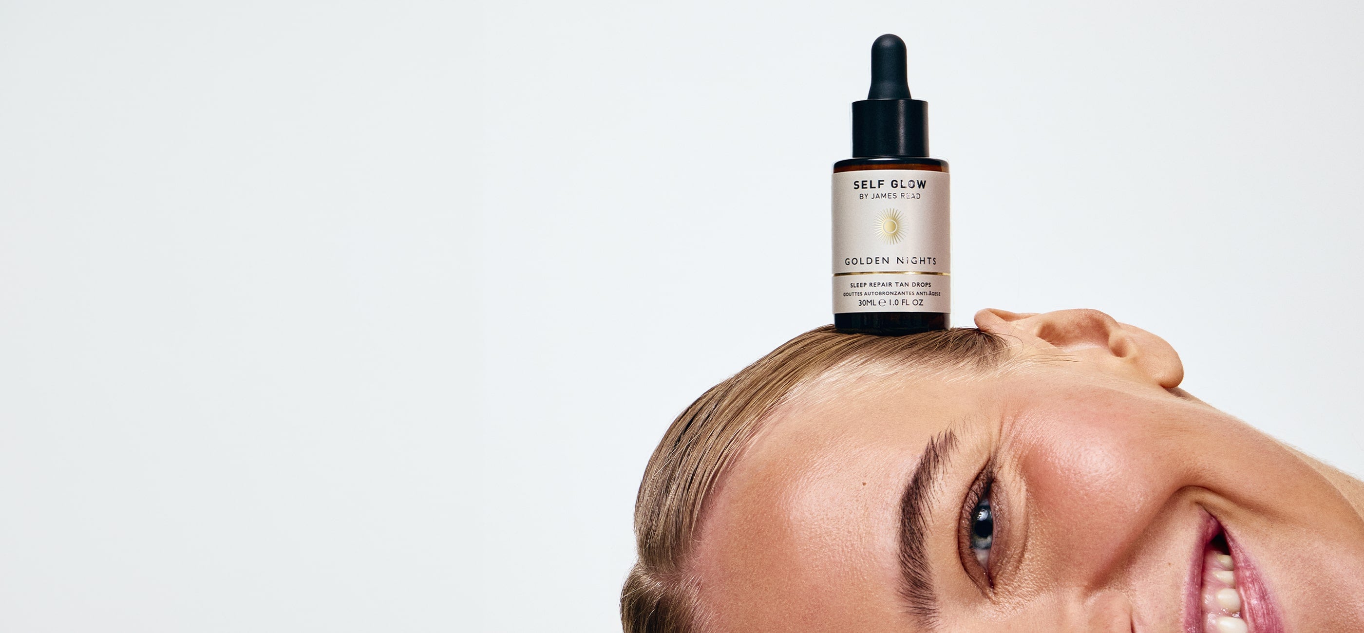 Model smiling with Self Glow by James Read Golden Nights Sleep Repair Tan Drops bottle balanced on head, showcasing natural glowing skin.