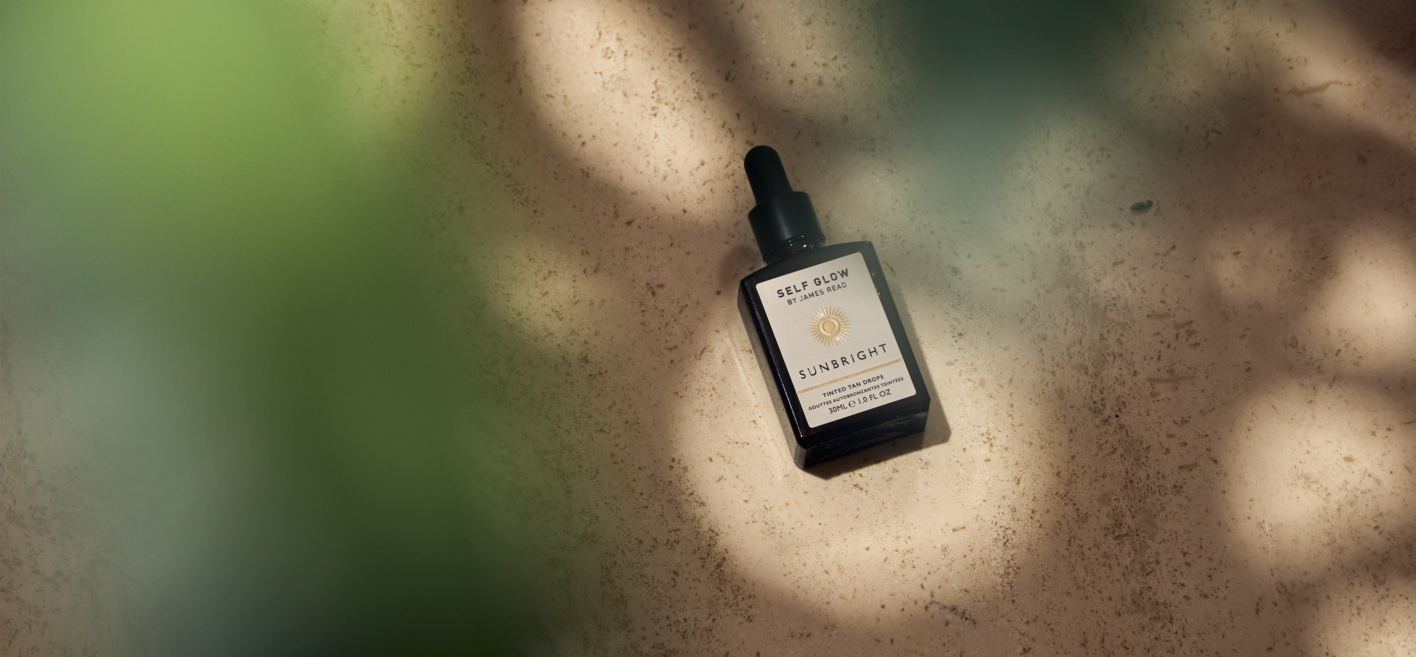 James Read's Self Glow Sunbright Tinted Tan Drops in a lifestyle image with dappled light through a leaf over the camera lens, showcasing the brand's commitment to using plant-based, clean ingredients that are kind to both the planet and the skin for a natural-looking fake tan.